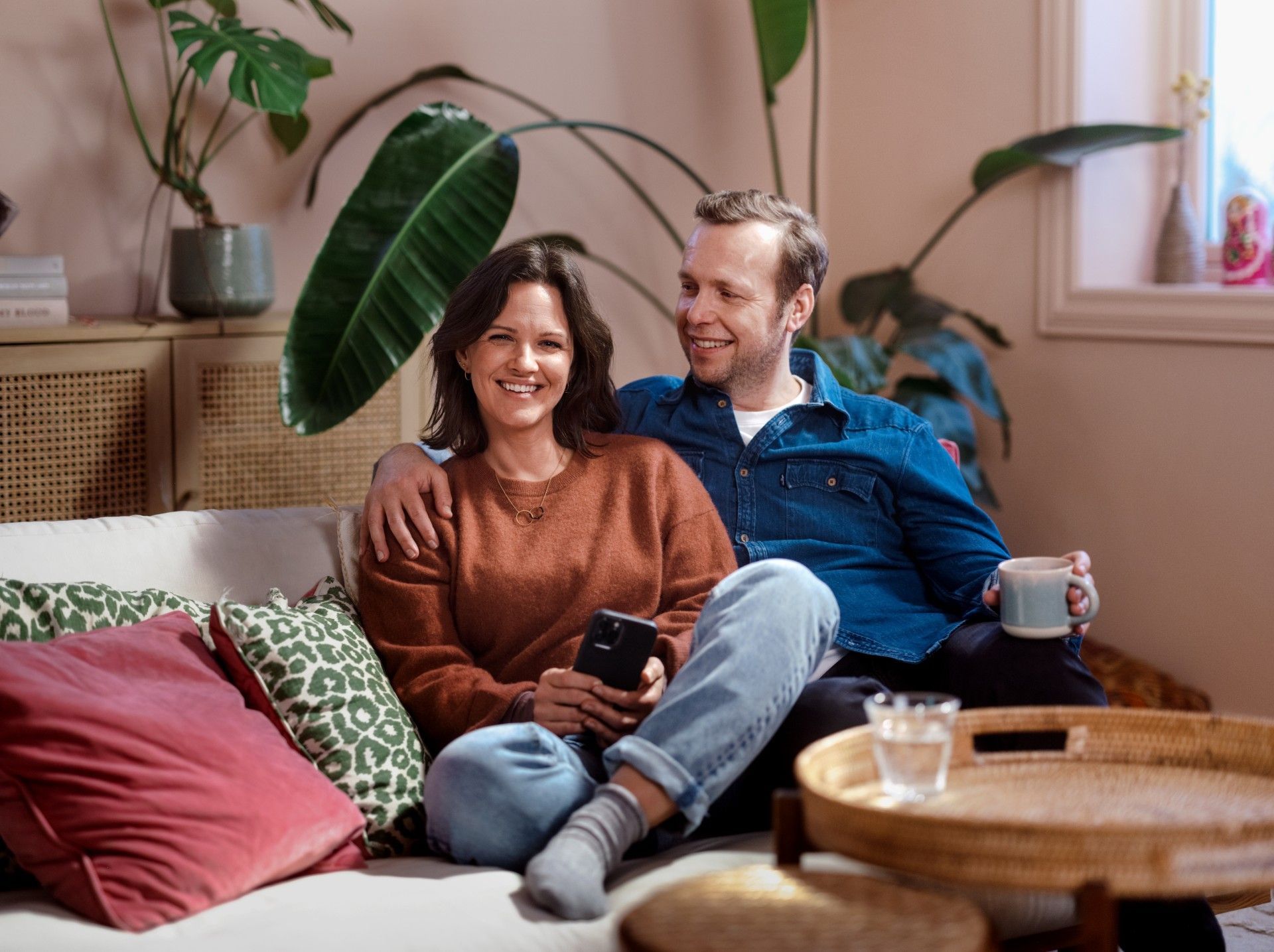Foto av kvinne og mann som sitter i en sofa. Han ser på henne og hun ser i kamera. I bakgrunnen er det møbler og planter.