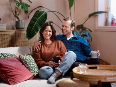 Foto av kvinne og mann som sitter i en sofa. Han ser på henne og hun ser i kamera. I bakgrunnen er det møbler og planter.