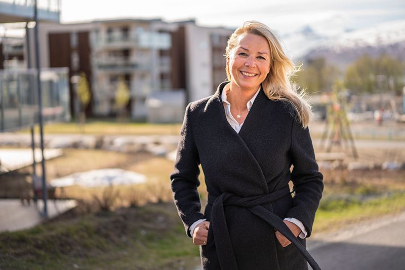 Dame står foran et boligblokkområde, smiler og ser inn i kamera. Bilde.