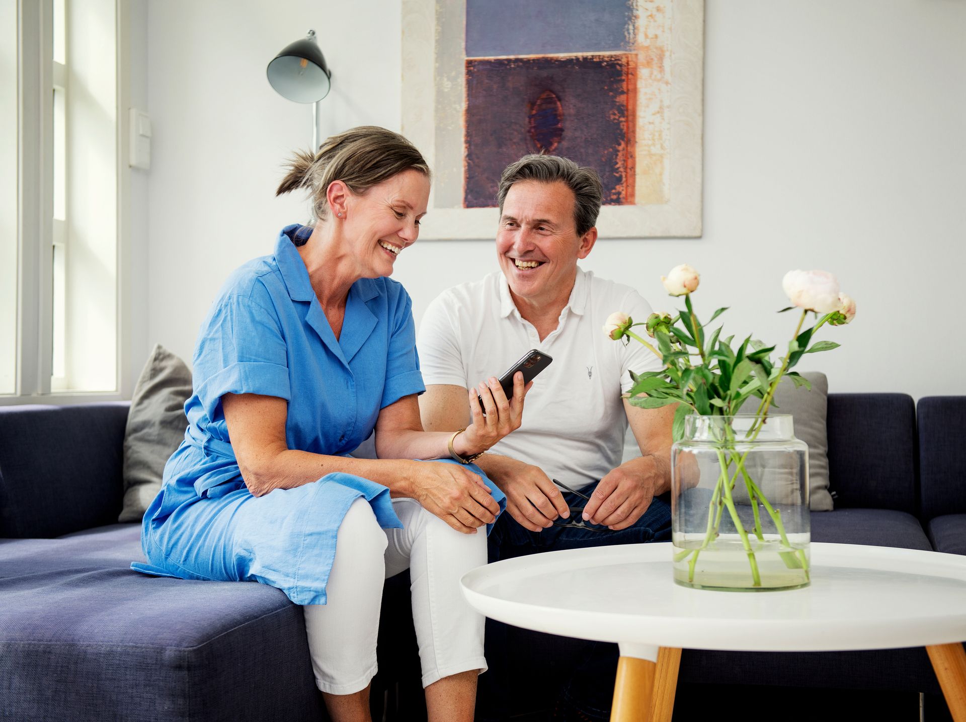 Foto av mann og kvinne som sitter sammen i en sofa. Begge smiler – han ser på henne og hun ser på en telefon.
