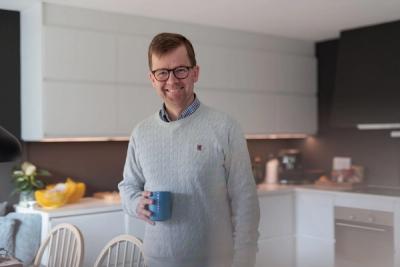 Foto av mann på et kjøkken. Han har en kopp i hånden og smiler til kameraet.