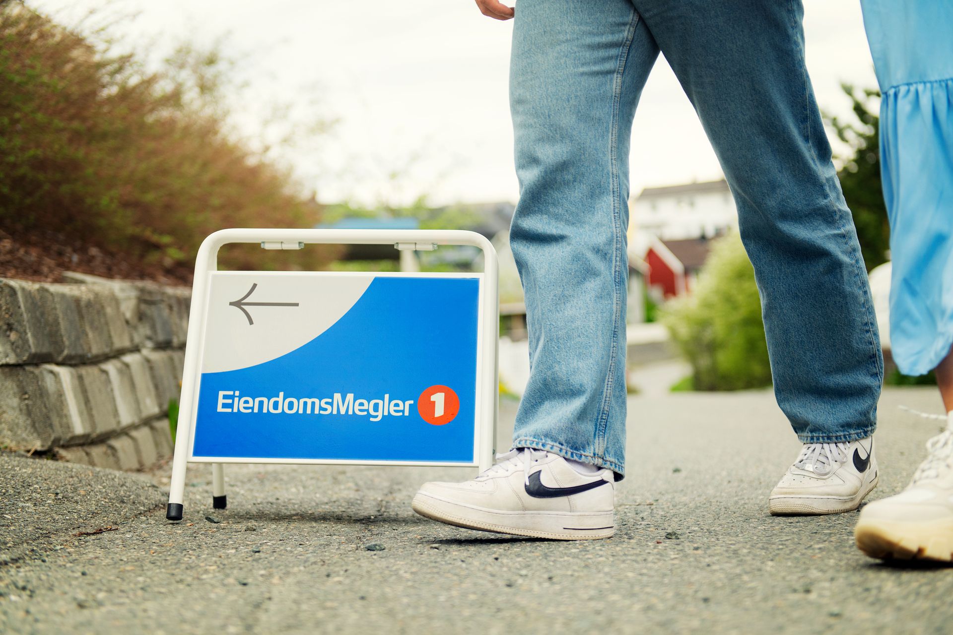 Foto av beina til to personer som passerer et visningsskilt med EiendomsMegler 1-logo.