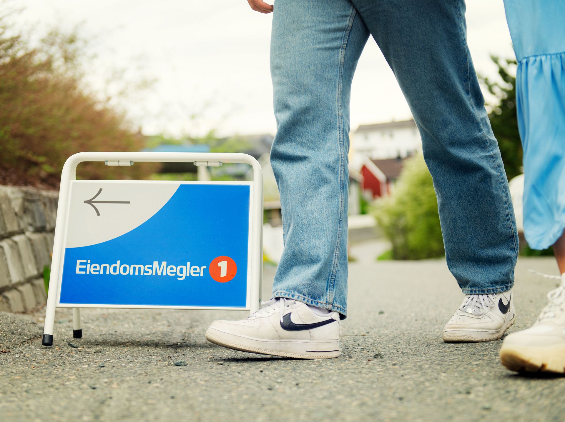 Beina til to personer passerer et visningsskilt med EiendomsMegler 1-logo. Foto.