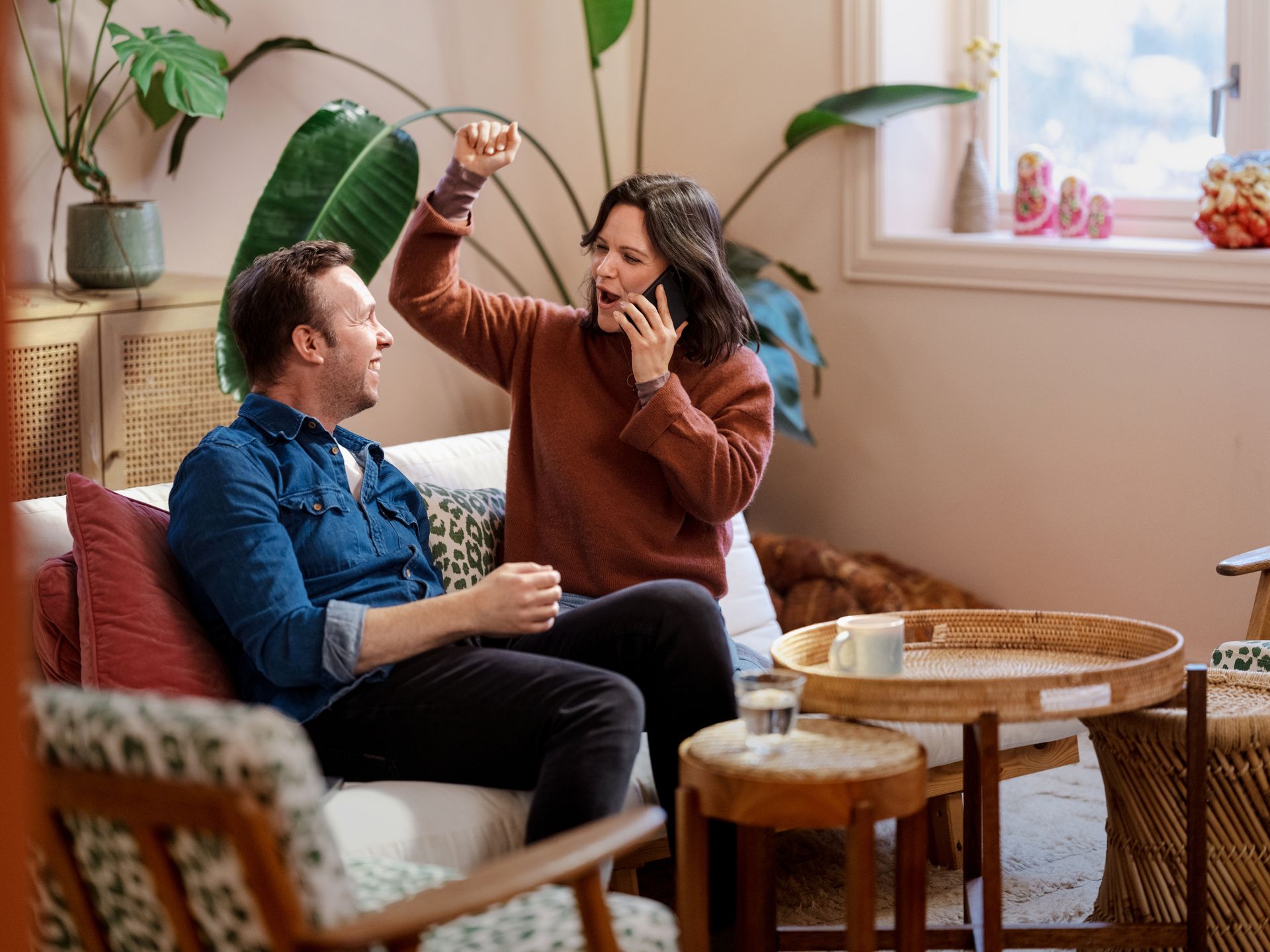 Par sitter i sofa. Kvinne snakker i telefon og jubler, mann ser på henne og smiler. Foto