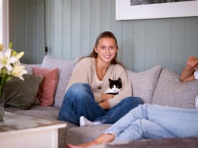 Foto av en ung kvinne som sitter i en sofa. Hun ser inn i kameraet, og i fanget holder hun en katt.