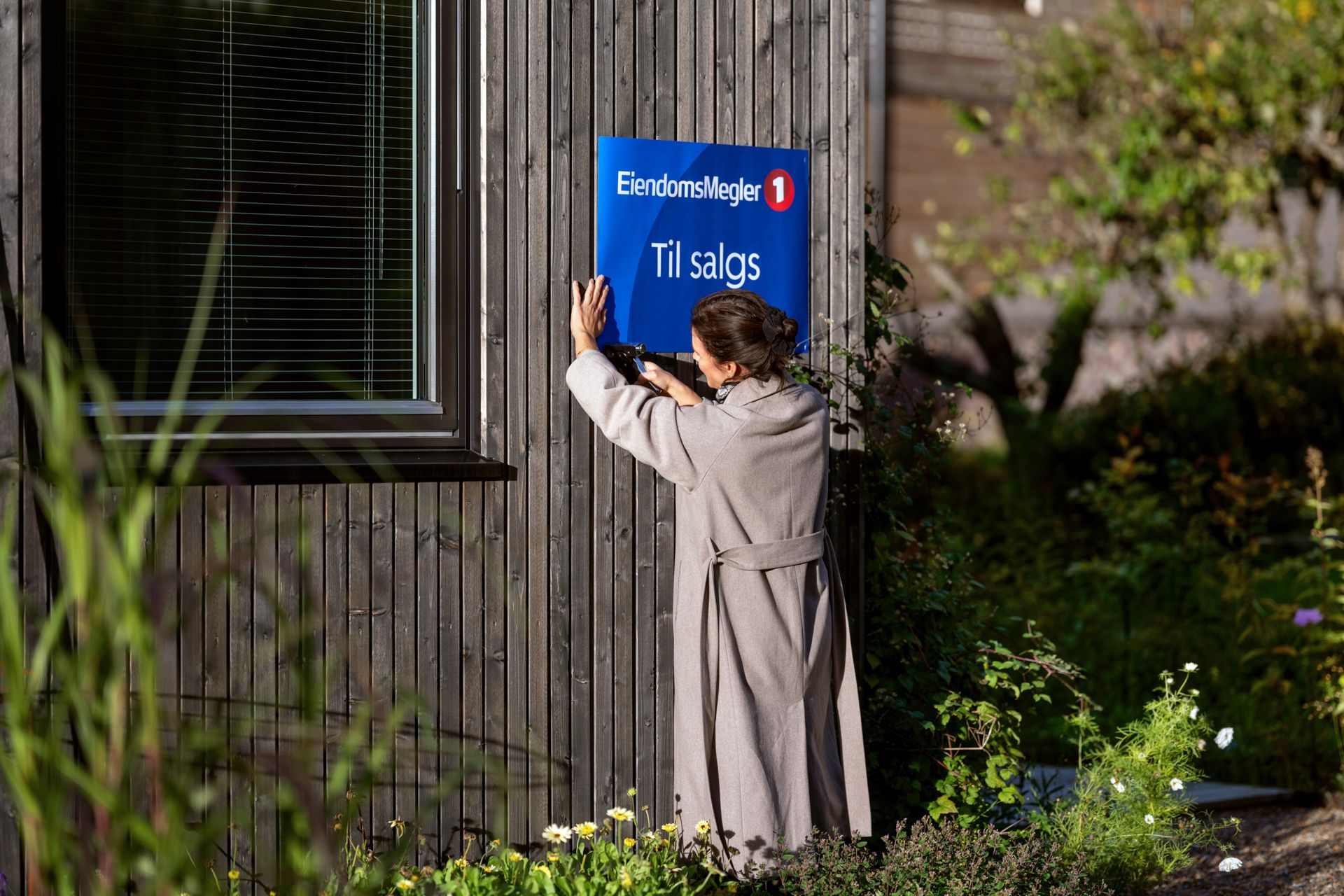 Dame henger opp en til salgs-plakat på en trefarget husvegg. Foto
