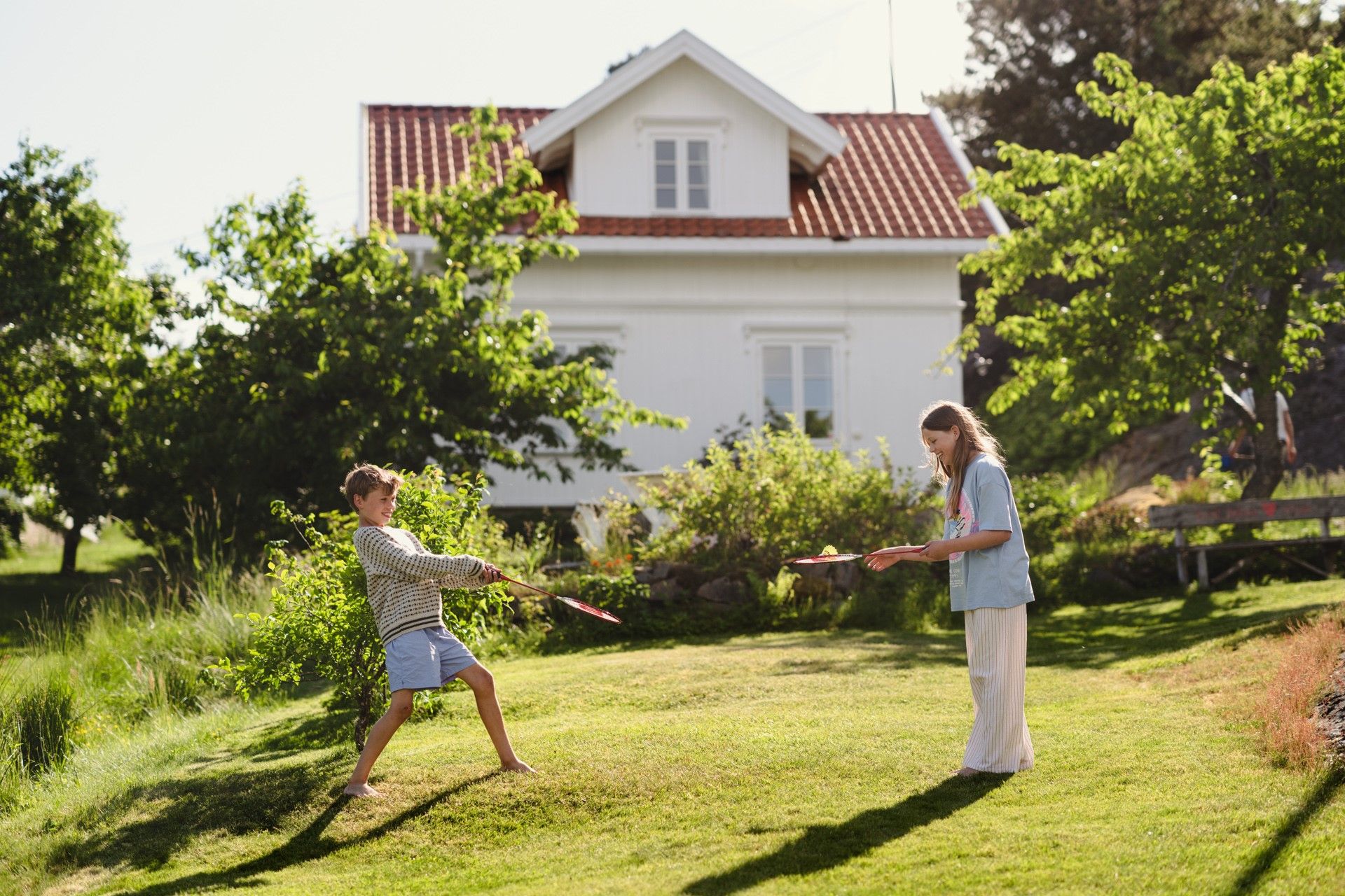 Jente og gutt spiller badminton. Hytte i bakgrunnen. Foto.