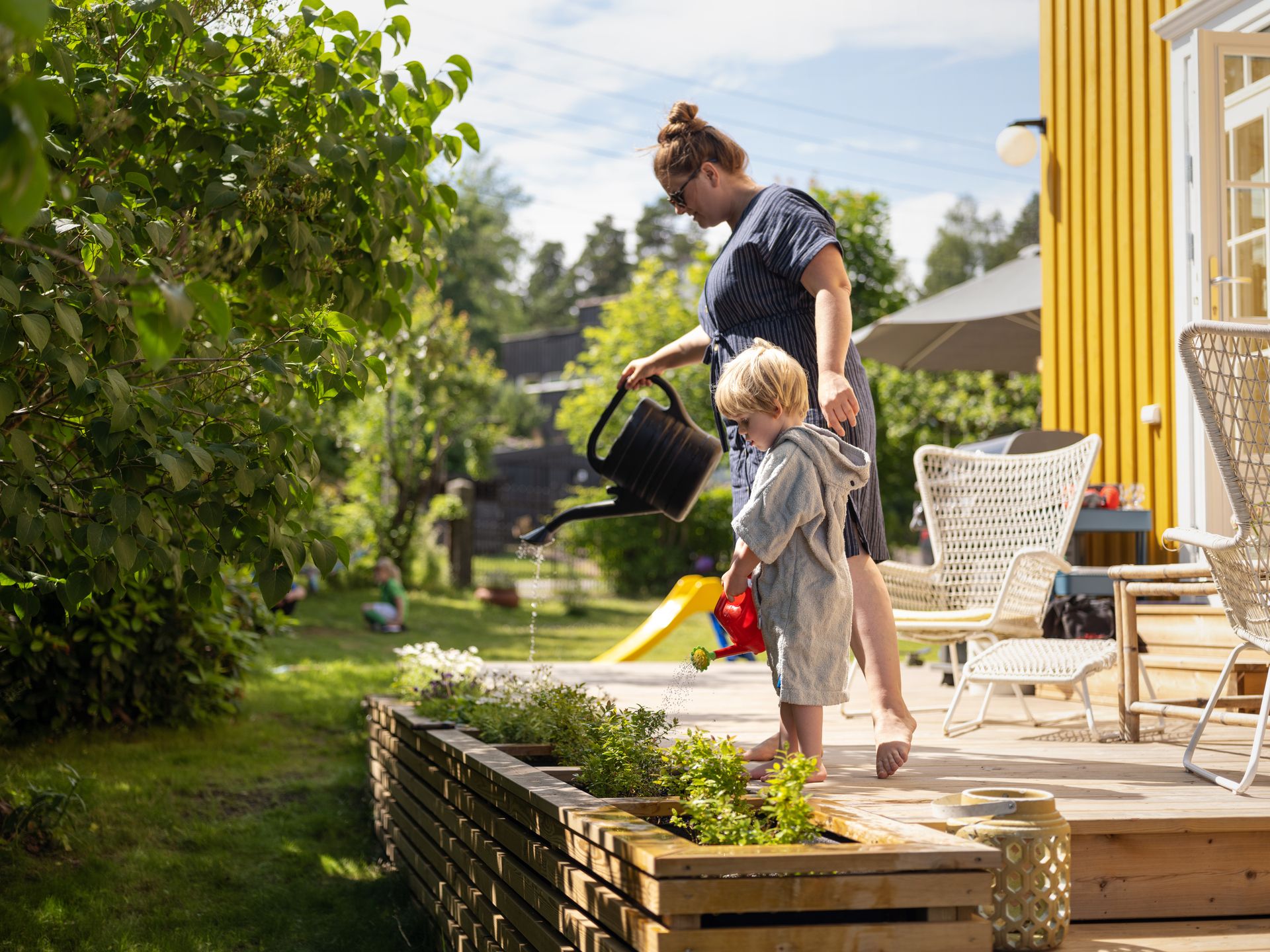 Foto av en dame og et barn foran et gult hus. De står på en terrasse og vanner grønne planter fra hver sin kanne.