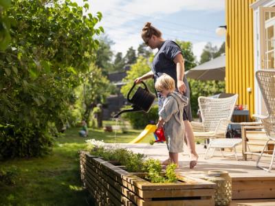 Foto av en dame og et barn foran et gult hus. De står på en terrasse og vanner grønne planter fra hver sin kanne.