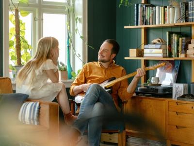 Foto av en pappa som spiller gitar for datteren sin. Han sitter i en stol, og hun på kanten av en sofa. De ser på hverandre