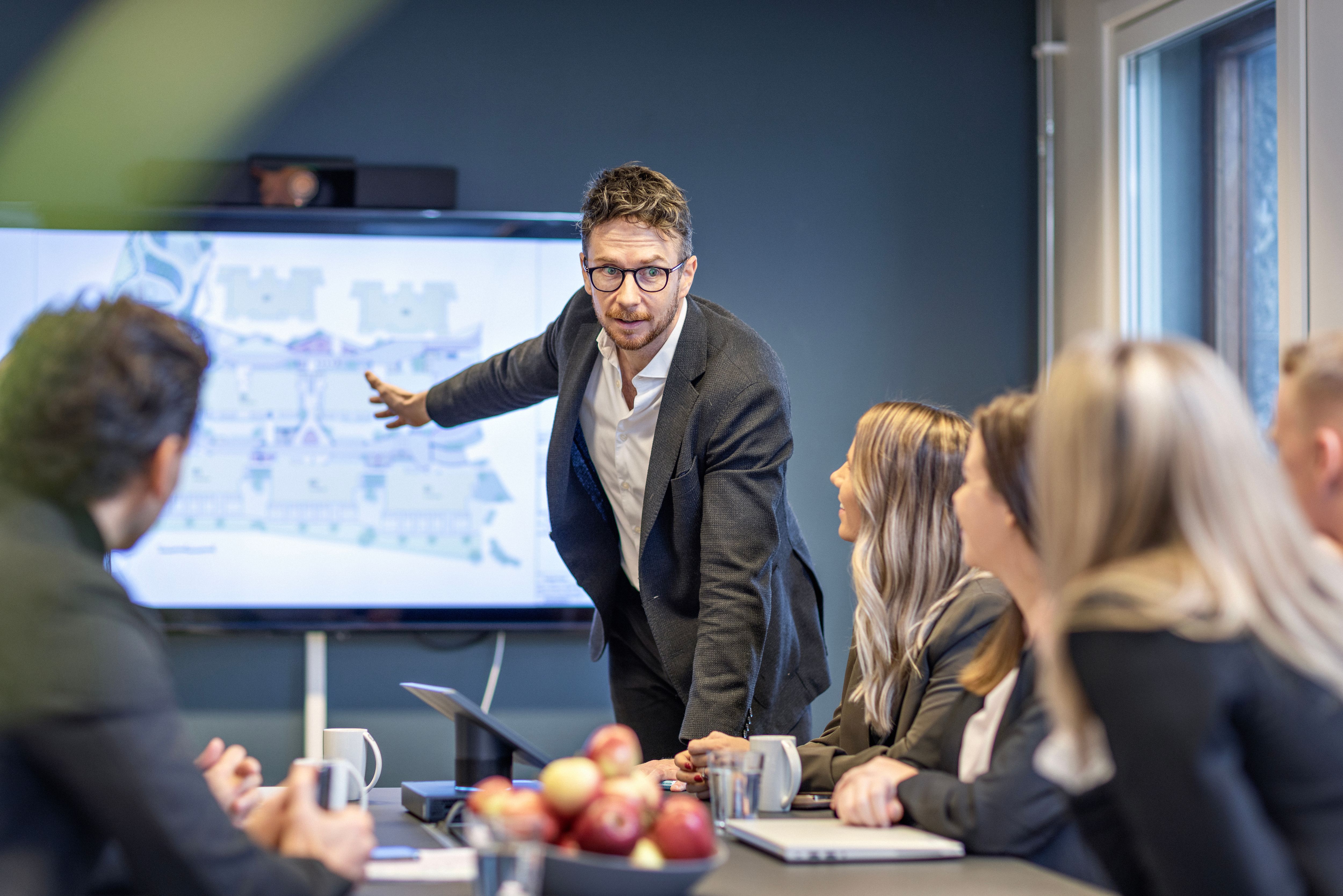 Næringsmegler holder presentasjon og peker på skjerm foran kollegaer.