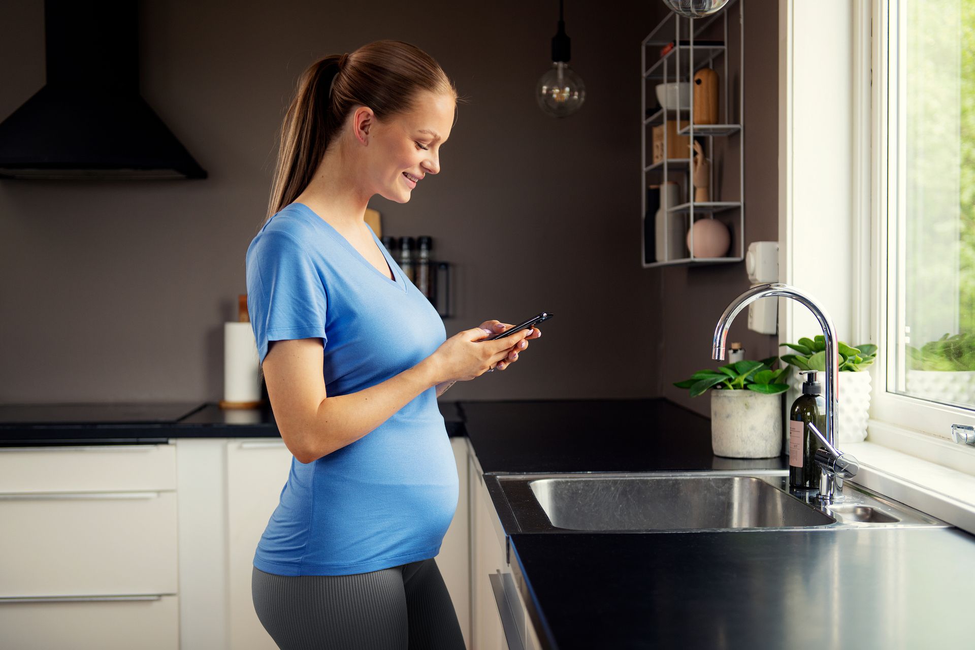Foto av gravid kvinne som står ved kjøkkenbenken og ser på mobilen.
