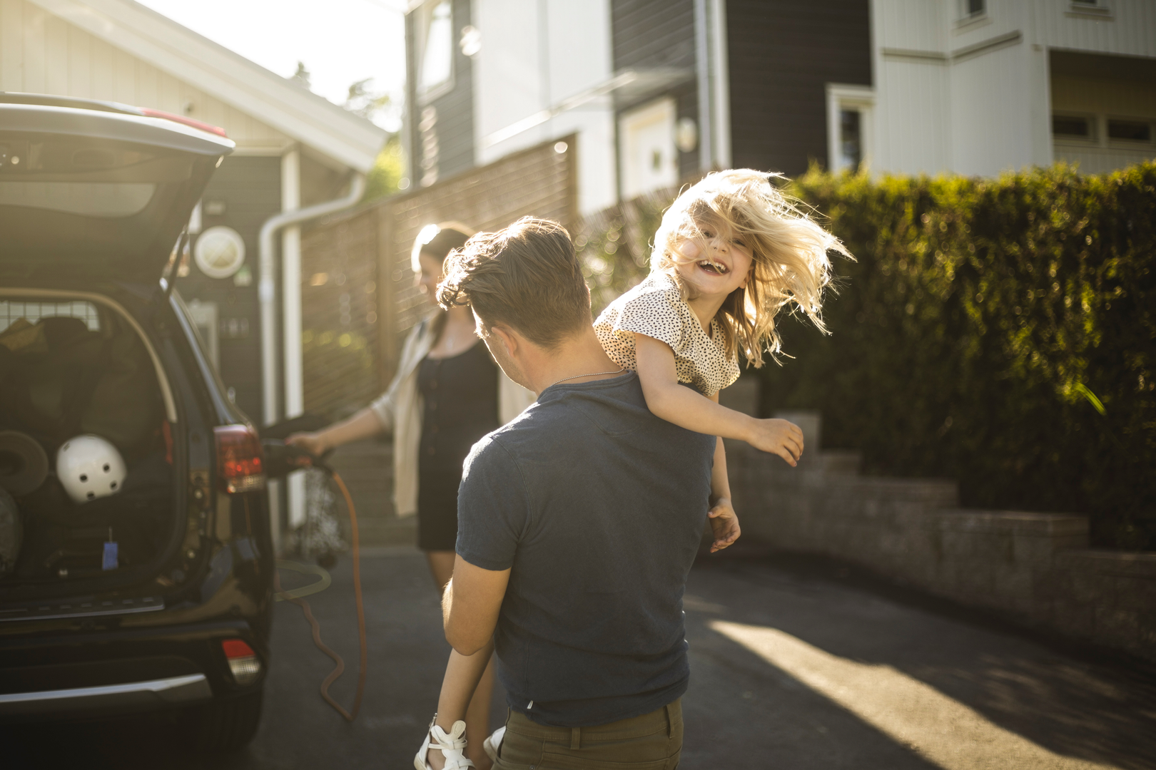 Foto av mann som bærer et smilende barn på skulderen. I bakgrunnen står mamma ved bilen og holder i ladeledningen.
