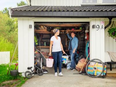 Par står i åpen garasjeport og snakker, kvinne bærer to bøtter. Foto.