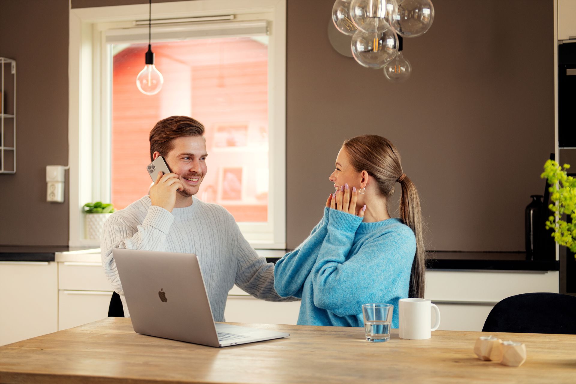 Foto av mann og kvinne som sitter ved et spisebord med en Mac oppå. Han smiler og snakker i telefonen, og kvinnen ser glad ut.
