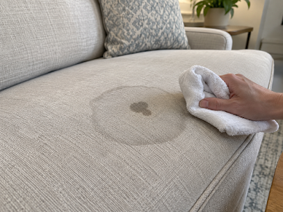 A hand blotting a ring-shaped stain on a light sofa cushion.