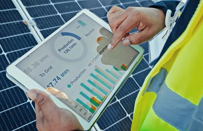 Eine Person in einer Warnweste benutzt ein Tablet, das Daten zur Solarenergieproduktion und -verbrauch vor Solarpaneelen anzeigt.