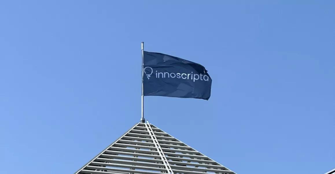 Dark blue flag with the white "innoscripta" logo waving atop a modern metal pyramid structure against a clear blue sky.