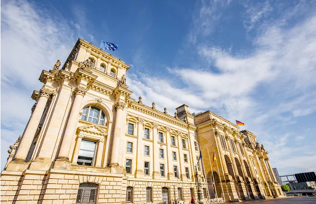 Wide-angle view of the historic Reichstag building in Berlin, Germany, with European Union and German flags flying under a bright blue sky with scattered clouds.
