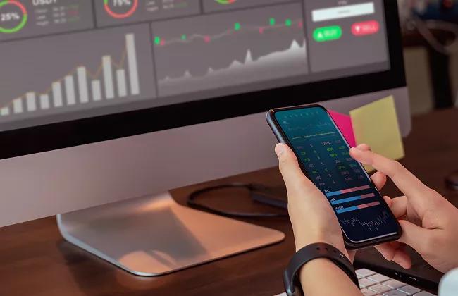 Person analyzing financial data on a smartphone with stock market charts displayed on a desktop monitor in the background.