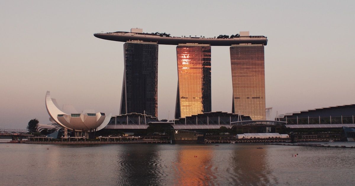 Three glass towers of Marina Bay Sands with rooftop skypark reflected in the bay at sunset, with the ArtScience Museum visible to the left.