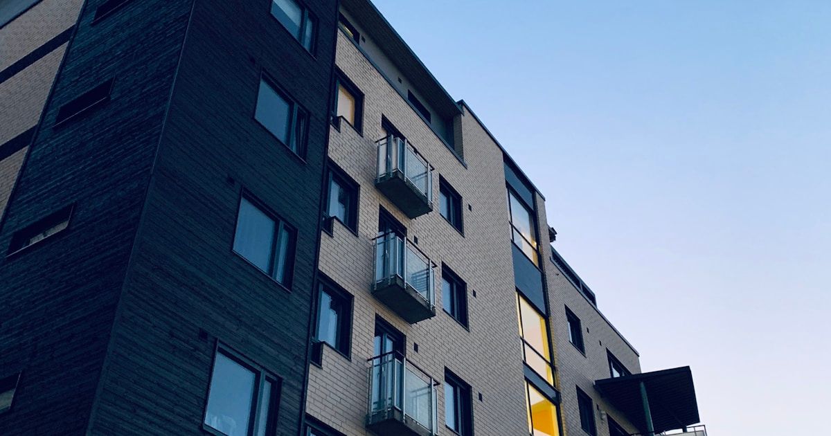 Low-angle view of a multi-storey brick apartment building with glass-railed balconies against a clear blue sky at dusk.