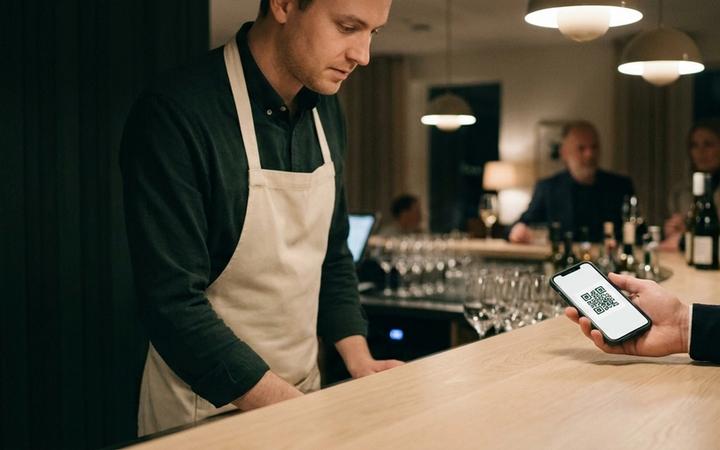 En gäst räcker fram sin mobil med ett digitalt presentkort som QR-kod över en bardisk i ljus ek, medan personalen lutar sig fram för att lösa in det.