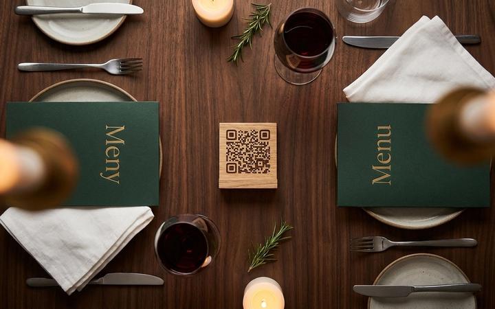 Bistrobord med en QR-kod på ett träställ och två menyer bredvid — "Meny" och "Menu" — i varmt kvällsljus