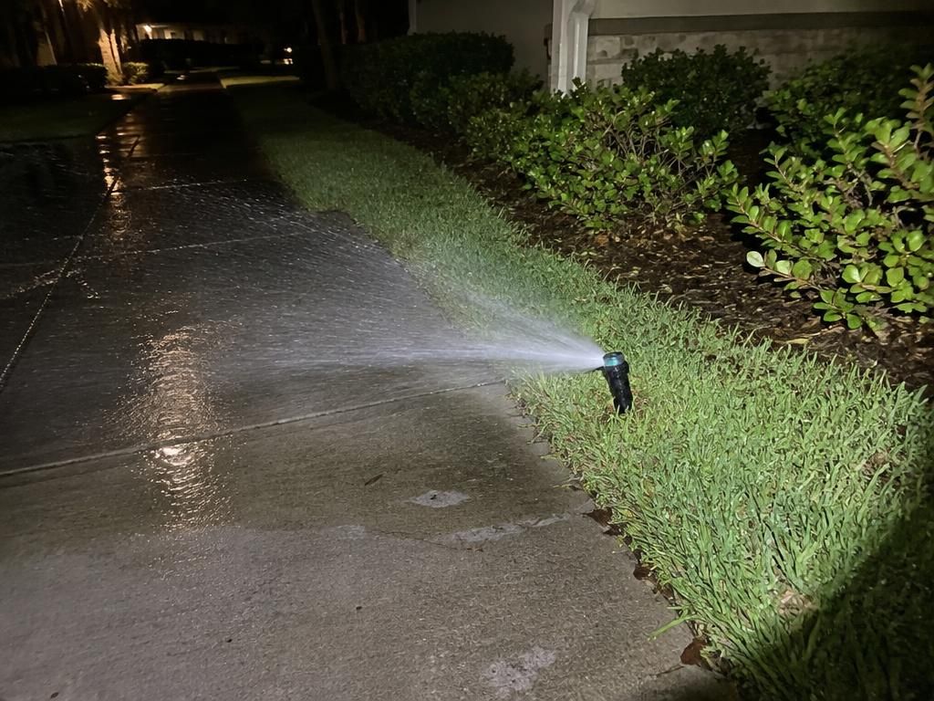 Sprinkler spraying water across a community sidewalk during the evening