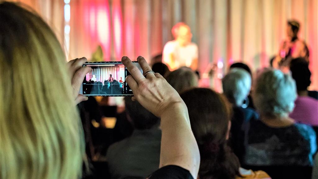 Bilde av arrangementlokale med samtale på scenen og en person tar bilde med mobilen.