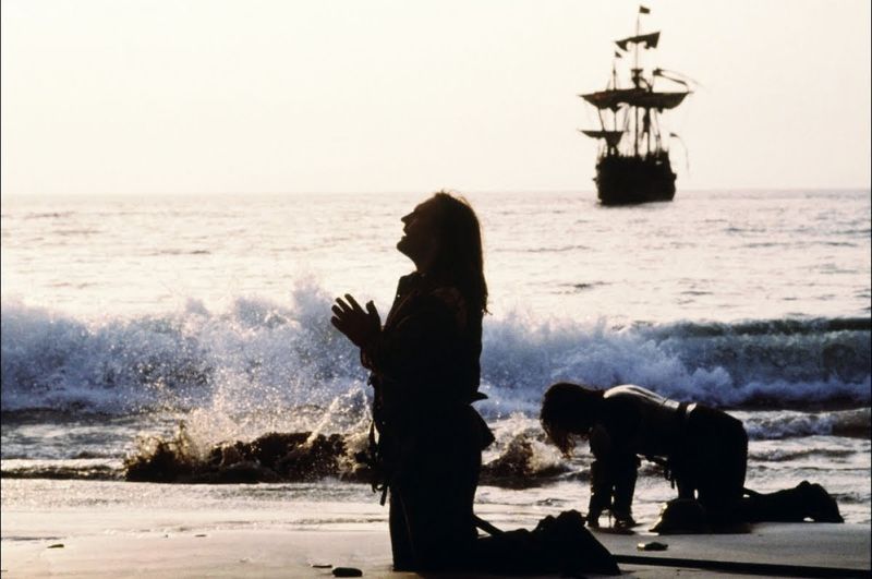 1492 Conquest of paradise film to menn kneler på strand med skip i bakgrunnen