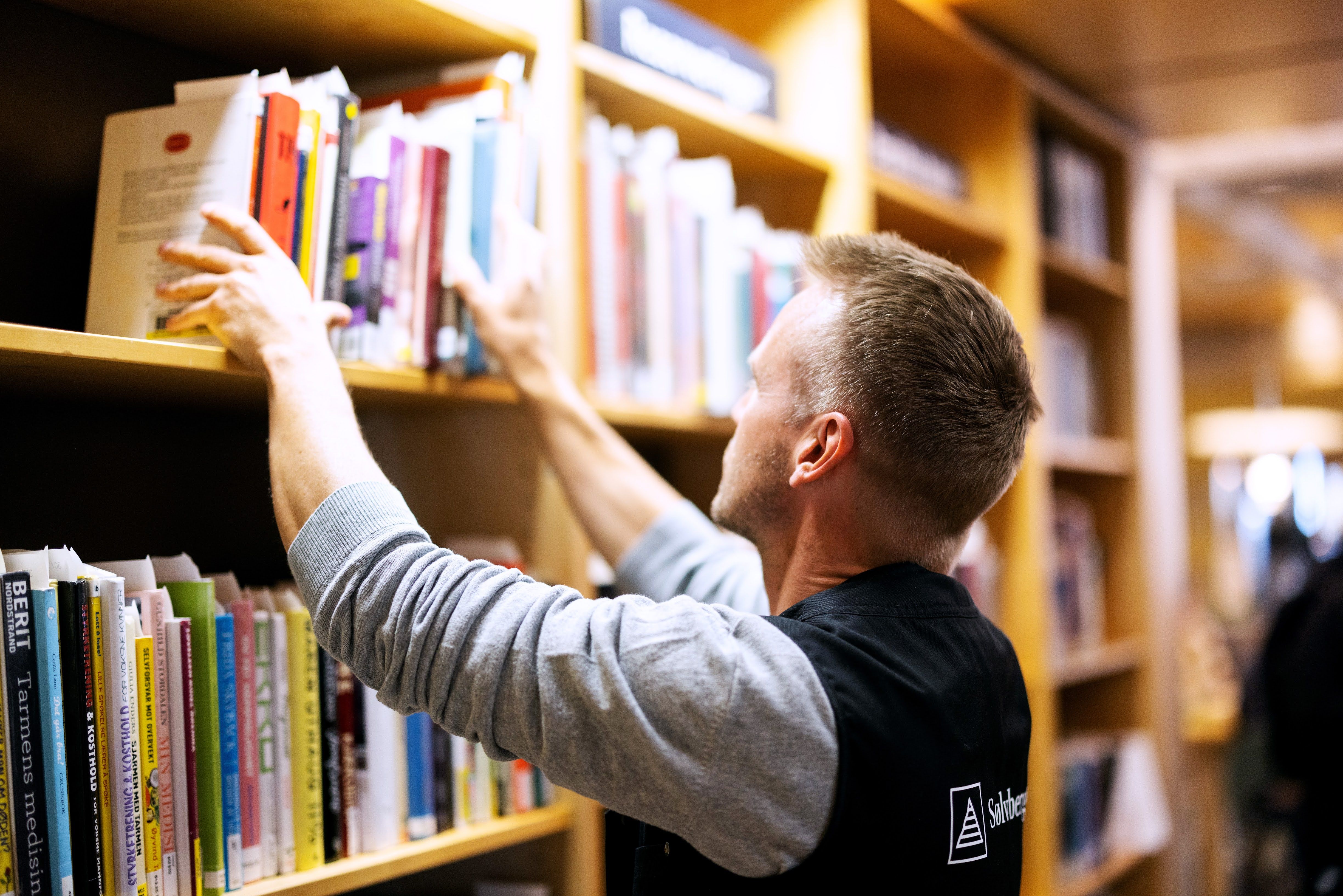 En ansatt med ryggen til setter på plass bøker i bokhylle.