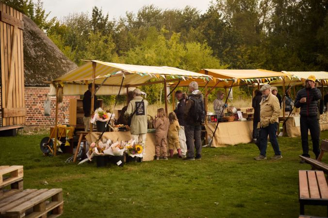 Gæster besøger tre af høstmarkedets boder, som er på en græsplads og omgivet af træer og et gammelt murstenshus med stråtag under en efterårshimmel.
