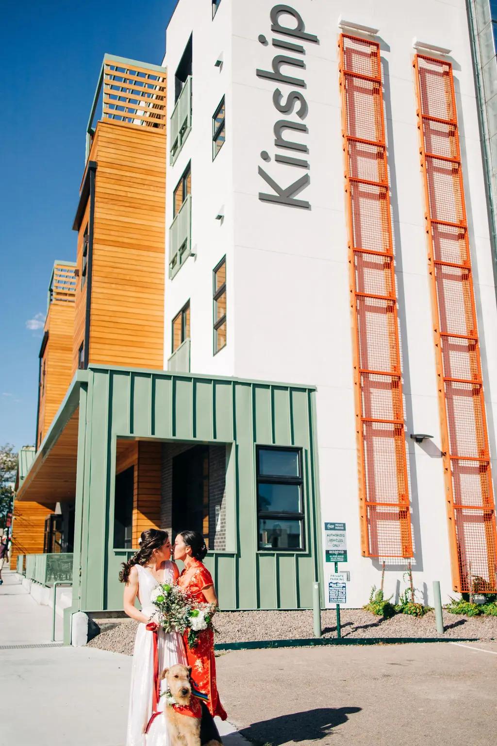 Two women in wedding attire and a dog standing in front of a building with the word 'Kinship' on it.
