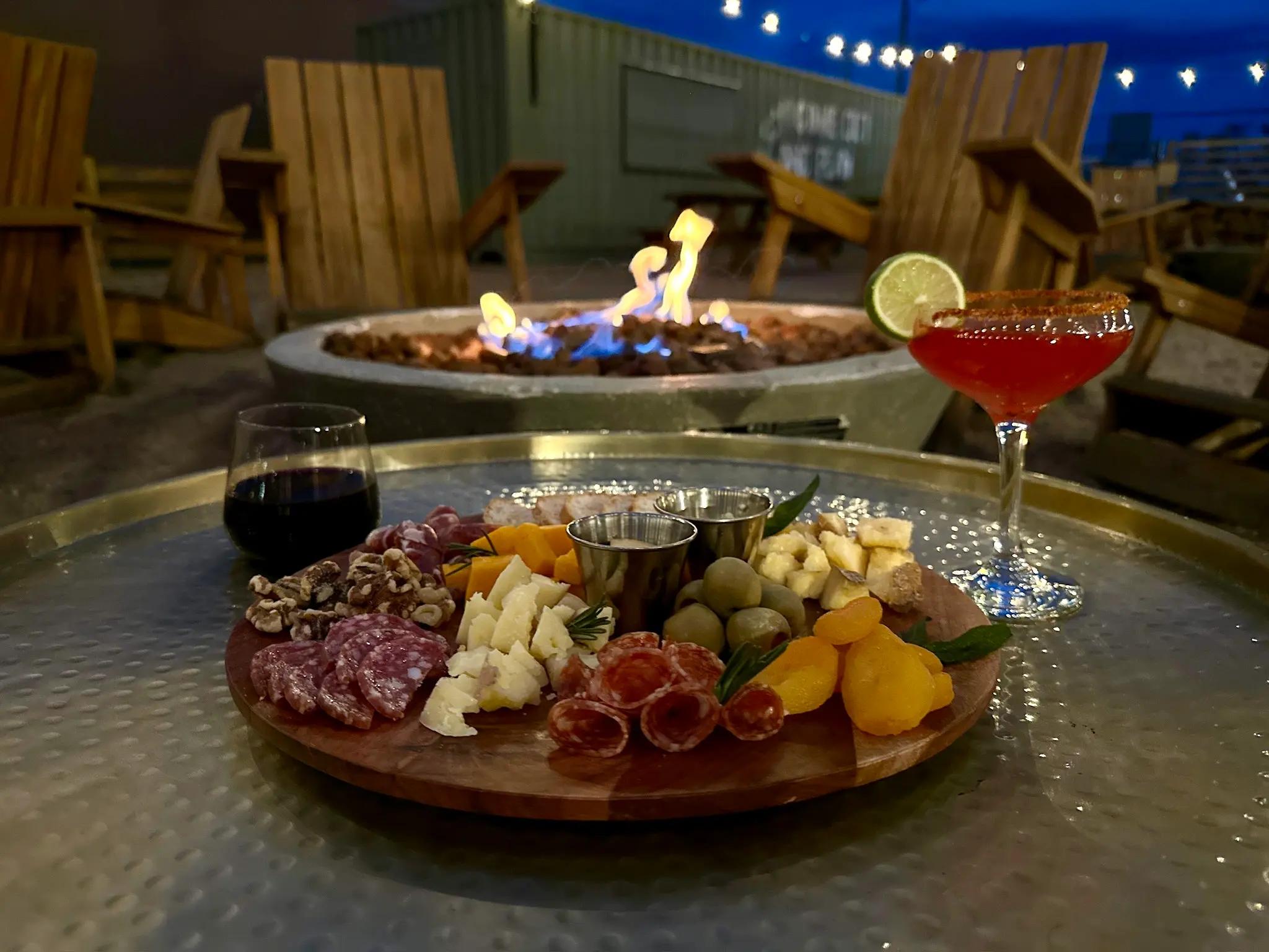 A charcuterie board with drinks in front of a fire pit.