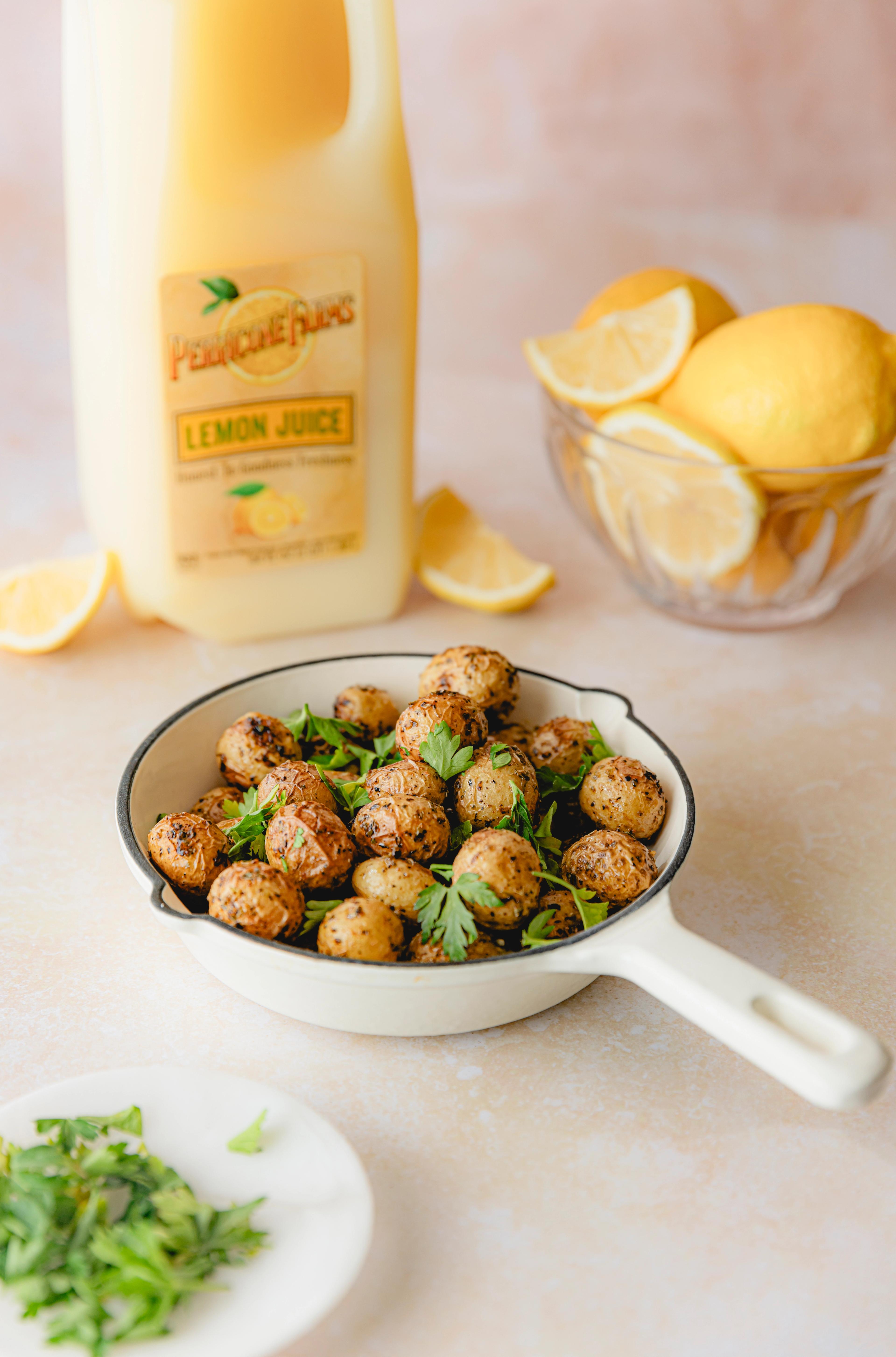 Roasted baby potatoes garnished with parsley, served in a white skillet next to a bottle of Perricone Farms Lemon Juice and a bowl of fresh lemons