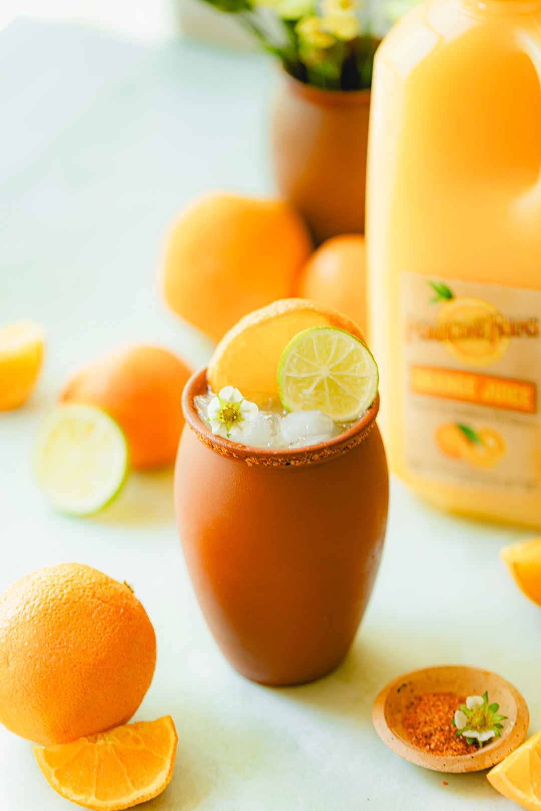 Classic Cantarito cocktail with Perricone Farms Orange Juice in a clay cup, Tajín rim, fresh citrus garnish, and whole oranges styled on a sunny tablescape.