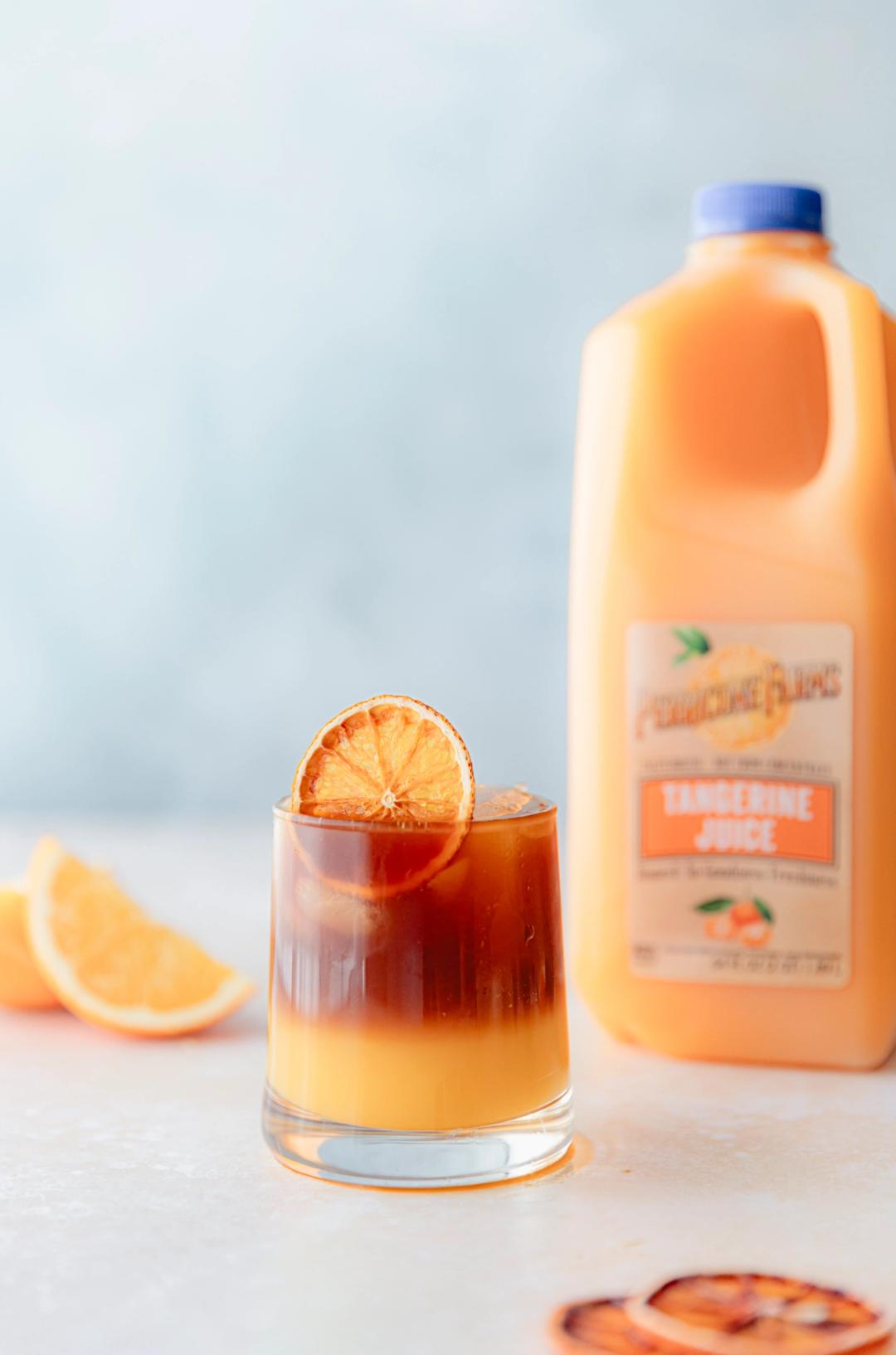 A layered tangerine espresso tonic in a clear glass with ice, topped with a dried orange slice beside a Perricone Farms Tangerine Juice jug.