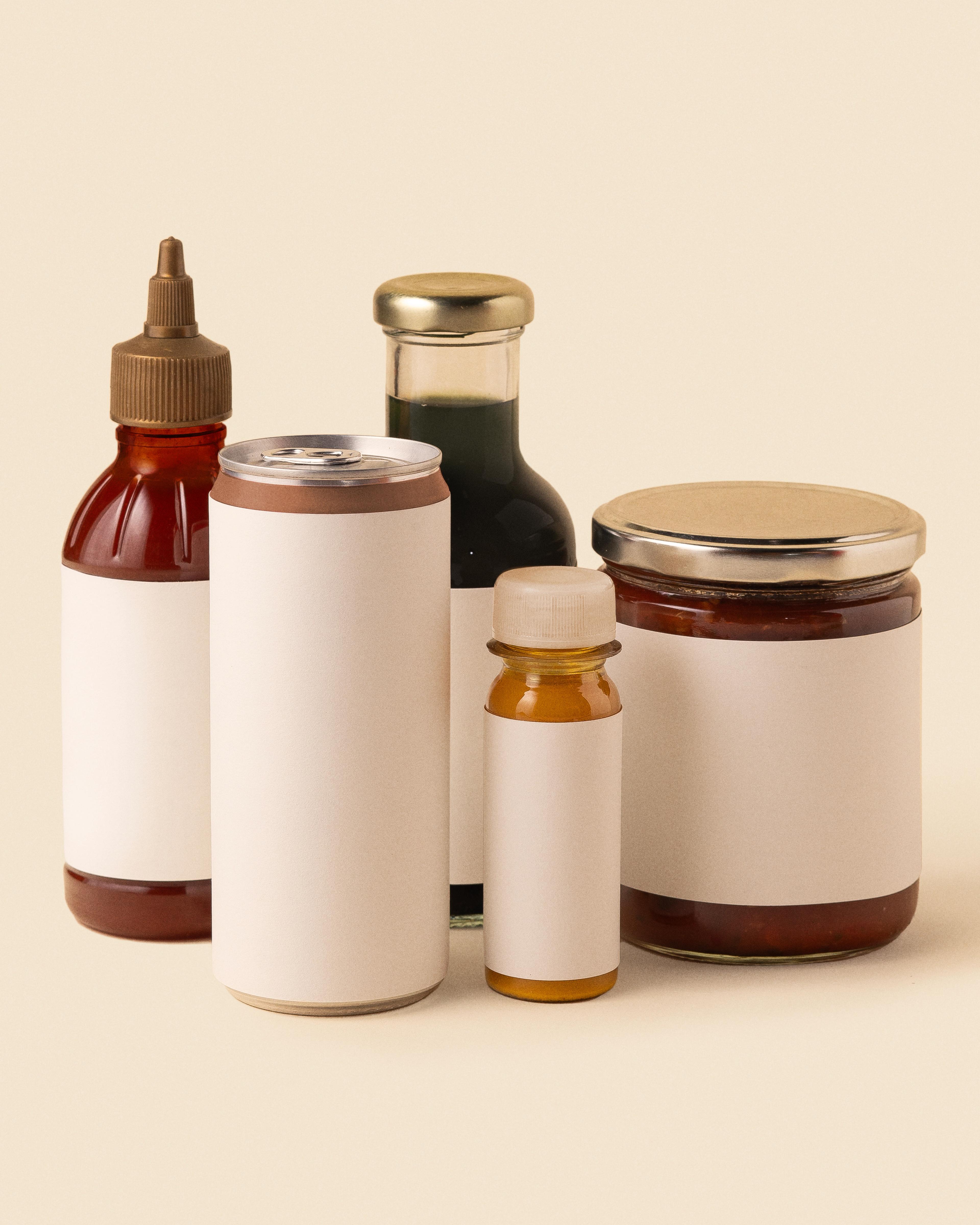 A group of food and beverage containers with blank white labels, including a squeeze bottle, aluminum can, glass bottle, plastic bottle, and glass jar, arranged against a neutral beige background.