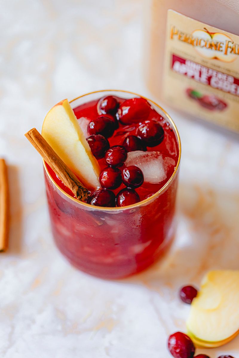 Cran-Apple Cider made with Perricone Farms Apple Cider and cranberry juice, served over ice with apple slices, frozen cranberries, and a cinnamon stick