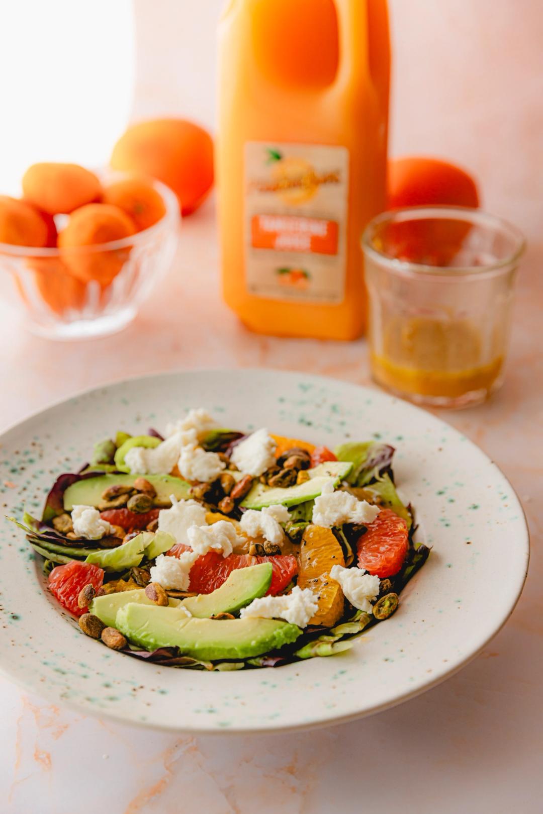 A colorful citrus salad topped with avocado slices, torn mozzarella, and pistachios sits in a white speckled bowl. A jug of Perricone Farms Tangerine Juice and a glass bowl of tangerines are in the background.