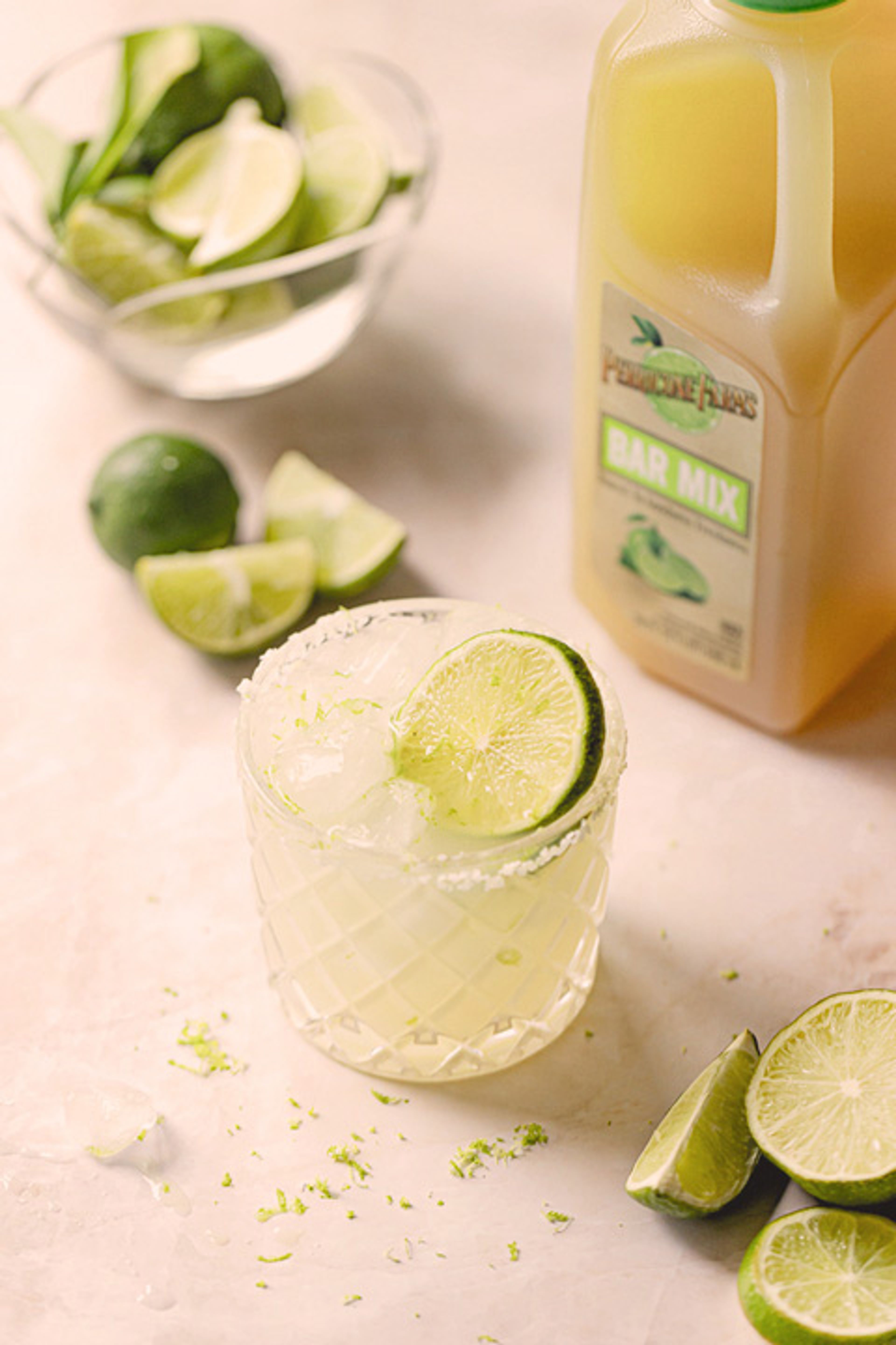 A margarita cocktail made with Perricone Farms Bar Mix served in a glass with a salted rim and lime garnish, surrounded by fresh lime wedges and a bottle of bar mix.