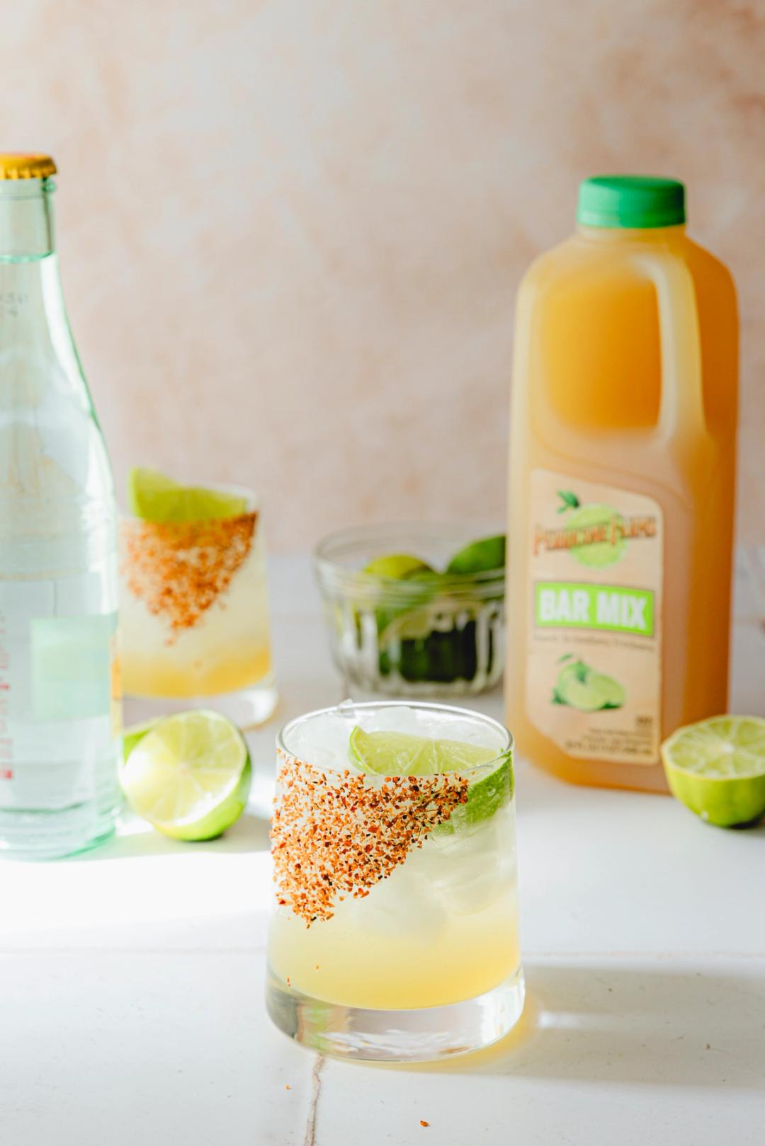 A chilled glass of mezcal ranch water with a Tajin-spiced rim and lime wedge garnish, served beside a bottle of Perricone Farms Bar Mix and halved limes on a white tile surface.
