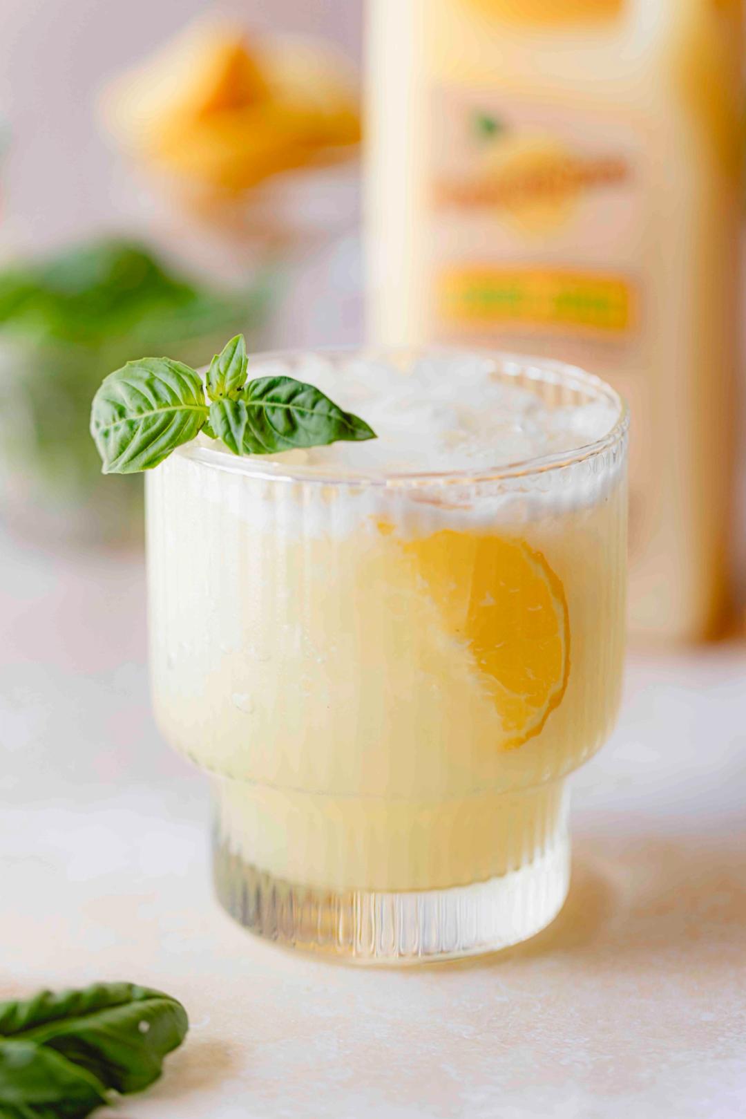 Lemon Basil Fizz made with Perricone Farms Lemon Juice, gin, and egg white served over ice with basil garnish beside a jug of lemon juice and fresh herbs