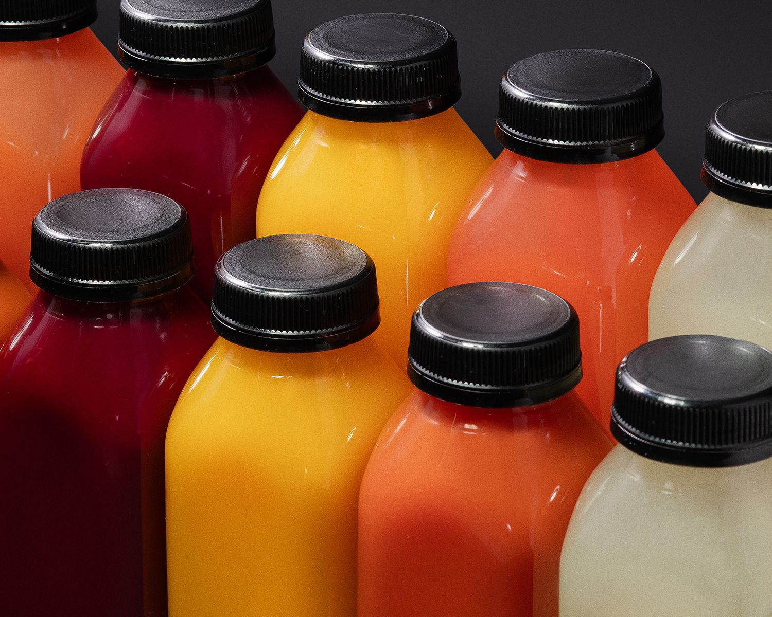 A close-up of colorful juice bottles with black caps, tightly arranged side by side in shades of deep red, golden yellow, and bright orange