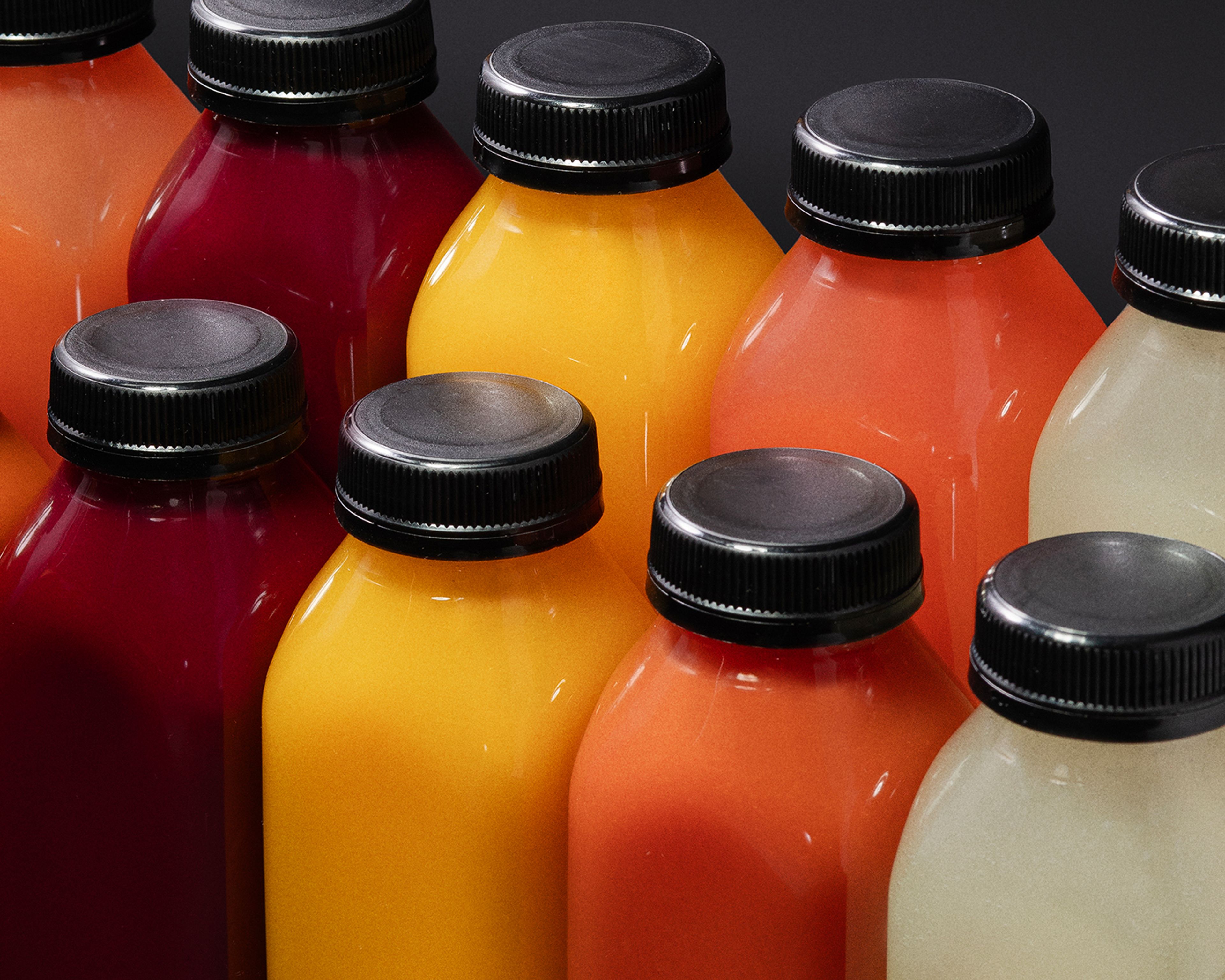 A close-up of colorful juice bottles with black caps, tightly arranged side by side in shades of deep red, golden yellow, and bright orange