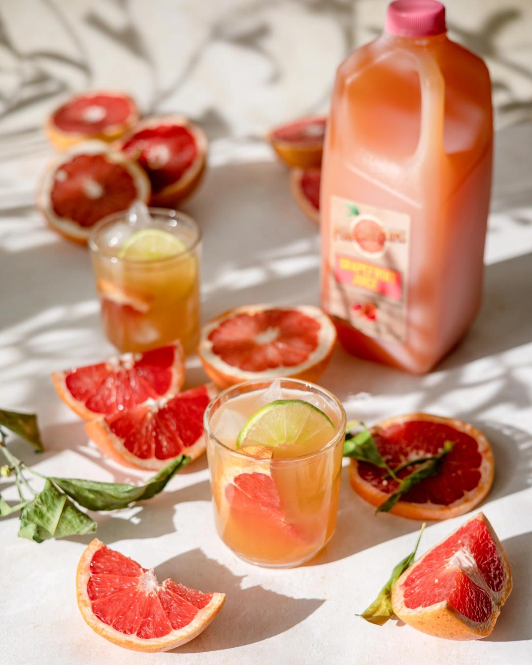 Aperol Paloma cocktail made with grapefruit juice, tequila, and lime, served over ice and garnished with a grapefruit slice and lime wheel. Surrounded by fresh grapefruit halves and a bottle of Perricone Farms grapefruit juice on a sunlit surface.