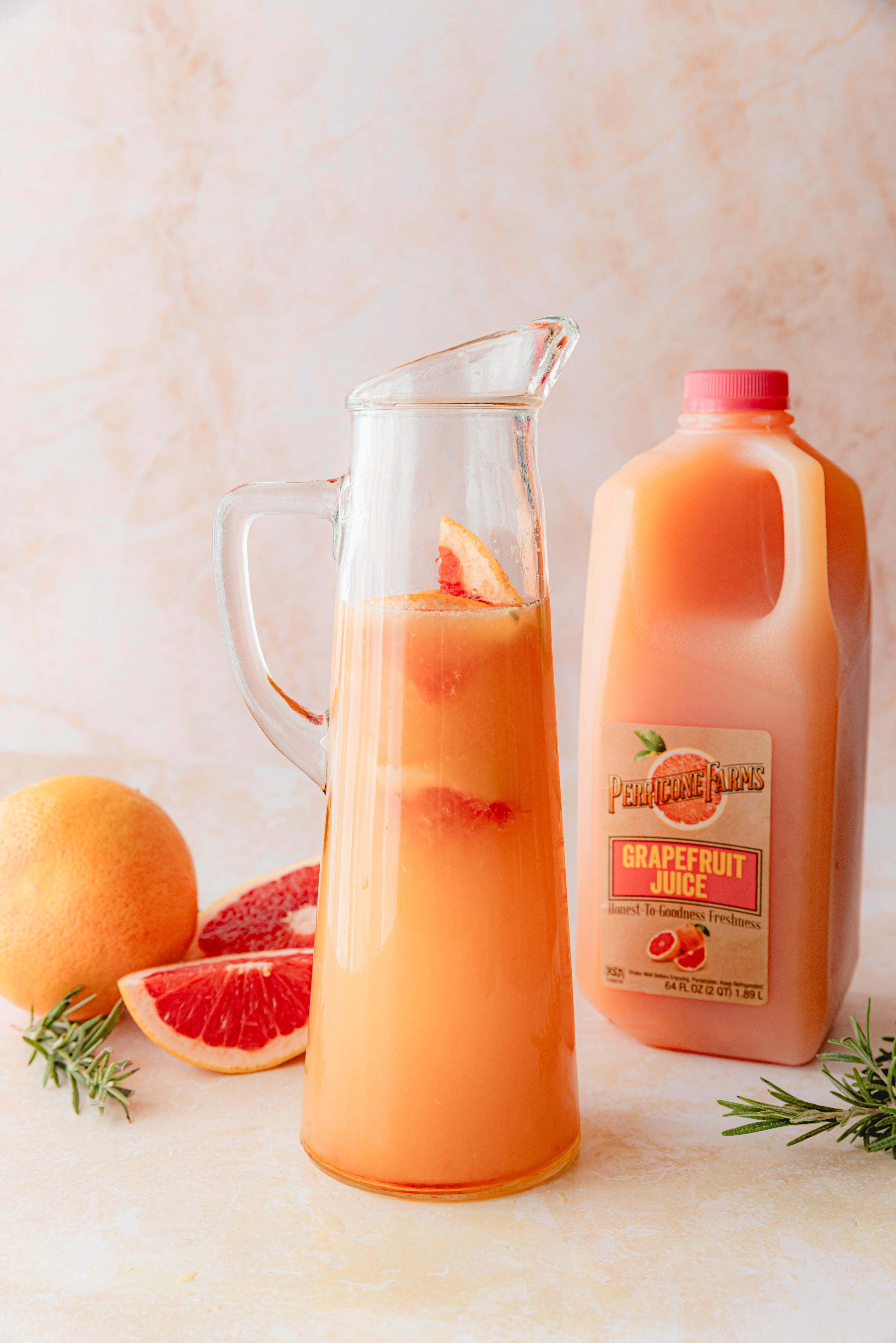 A refreshing grapefruit Paloma in a glass pitcher, garnished with rosemary and grapefruit slices. Perricone Farms Grapefruit Juice is visible in the background.