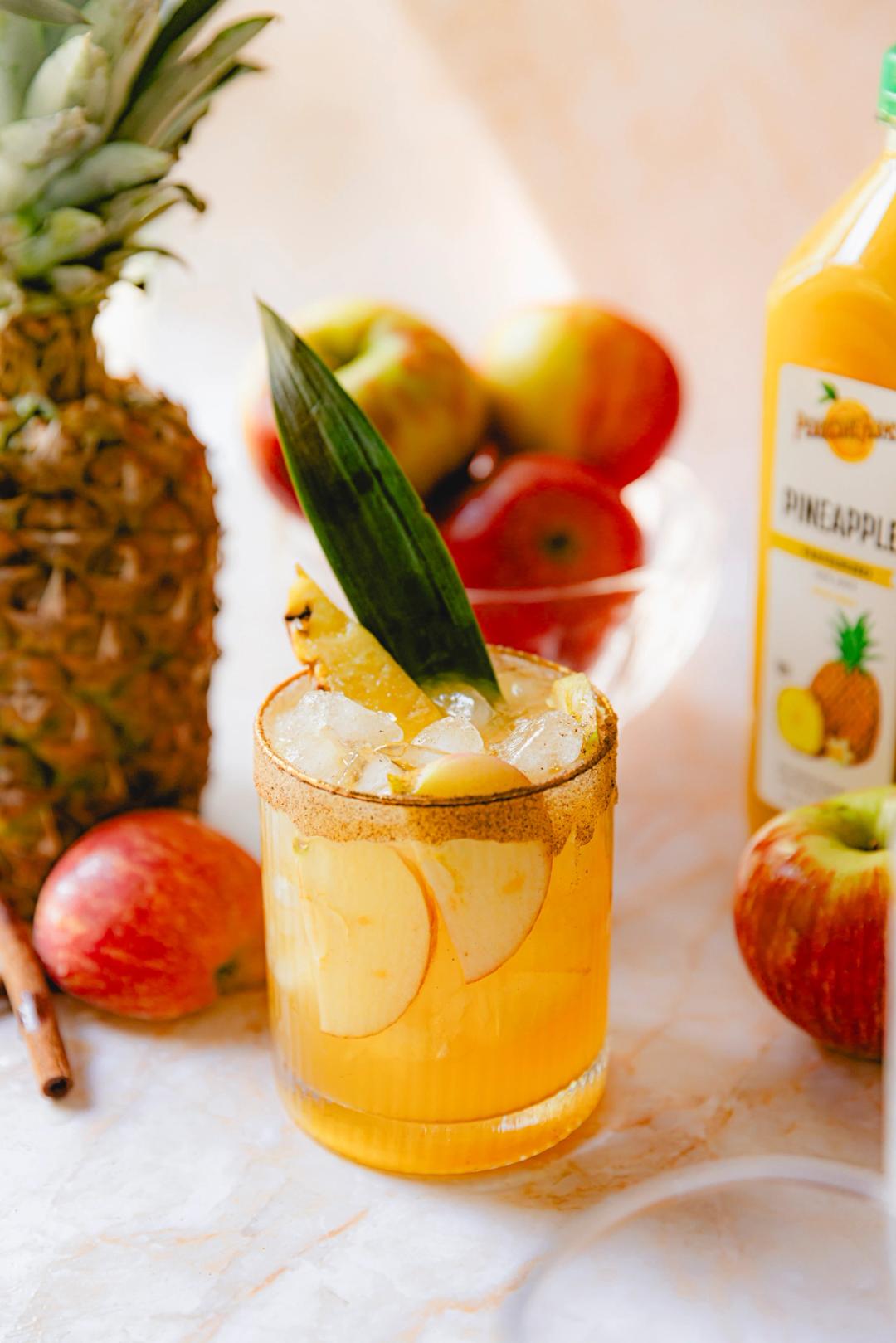 A spiced pineapple mocktail served in a short glass rimmed with cinnamon sugar. The drink is golden yellow and filled with ice, thin apple slices, and garnished with a pineapple spear and a pineapple leaf. Fresh apples and a whole pineapple are placed in the background next to a bottle of Perricone Farms Pineapple Juice.