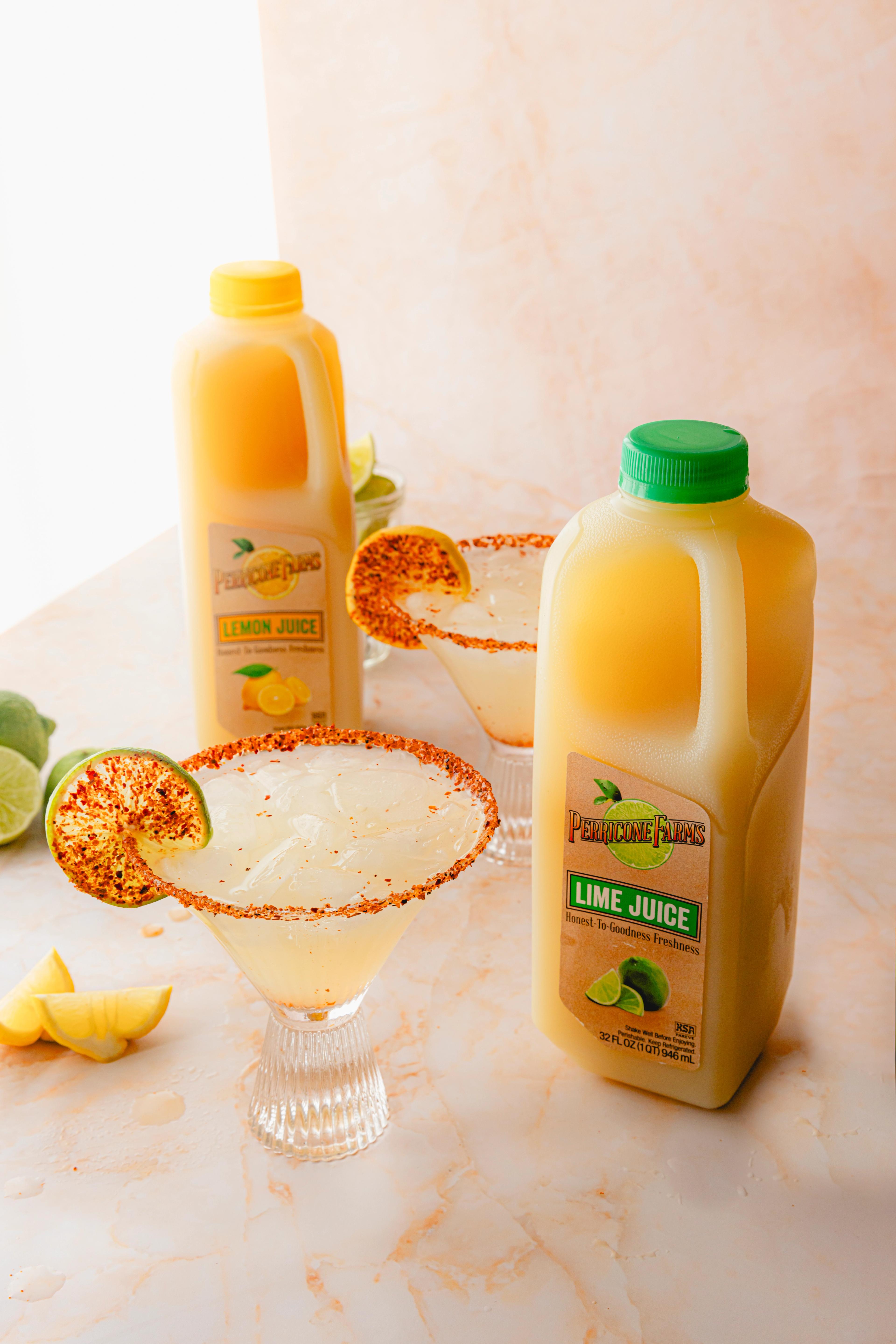 A chilled margarita served in a stemmed glass with a Tajín-spiced rim and garnished with a lime wheel. The drink is pale golden and surrounded by fresh citrus and Perricone Farms lemon and lime juice bottles.